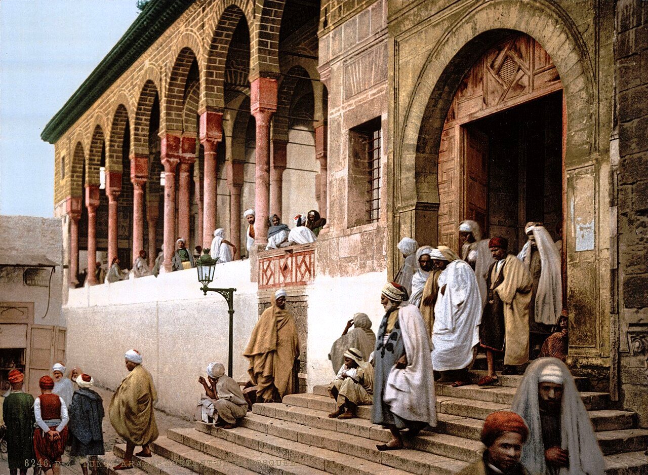 Men in long robes and turbans sit and stand on broad stone steps before an arched doorway and a colonnade in Tunis.