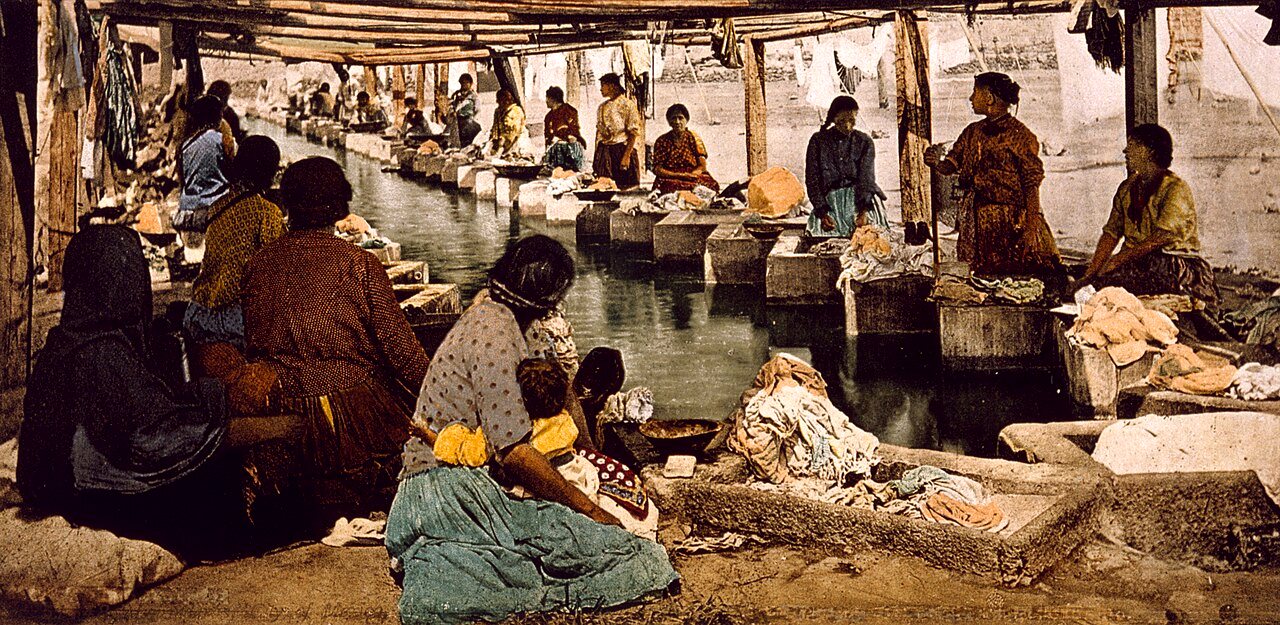 Women wash clothes along a water channel under a covered shed, with piles of laundry around them.
