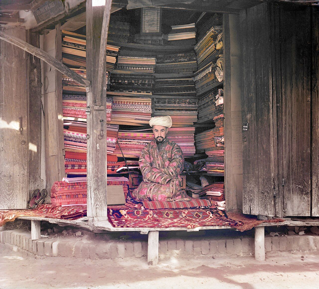 A man in a turban and a patterned robe sits cross-legged on carpets in a wooden stall, with tall stacks of folded fabrics rising behind him almost to the roof.