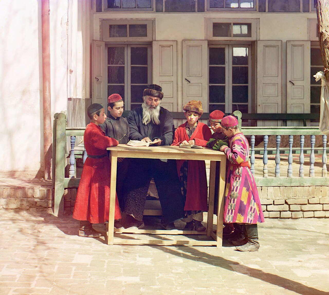 A white-bearded teacher in a dark coat and fur-trimmed hat reads from an open book at a wooden table, with five boys in red robes, caps and one bright patterned coat gathered around him in a sunlit courtyard.