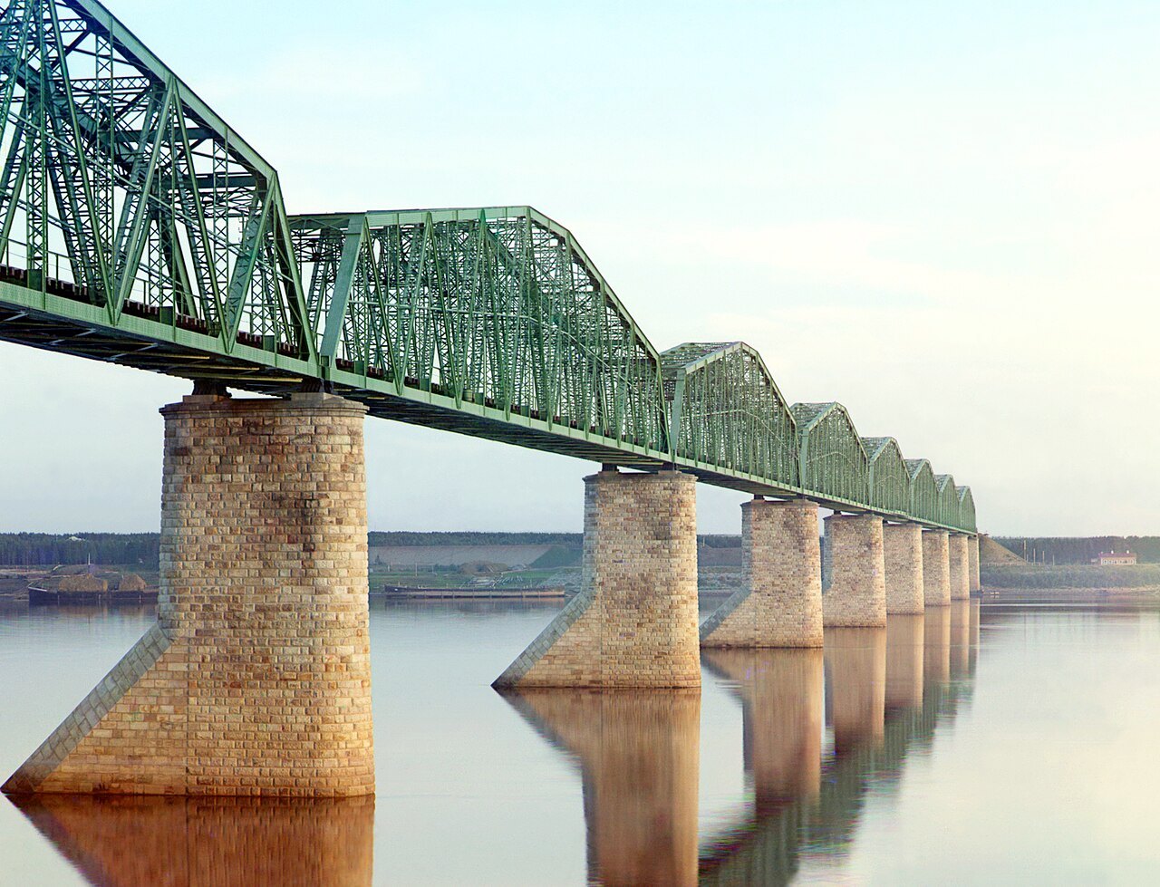 A long railroad bridge with green metal trusses crosses a wide, calm river on tall stone piers that shrink into the distance, their reflections lying in the still water.