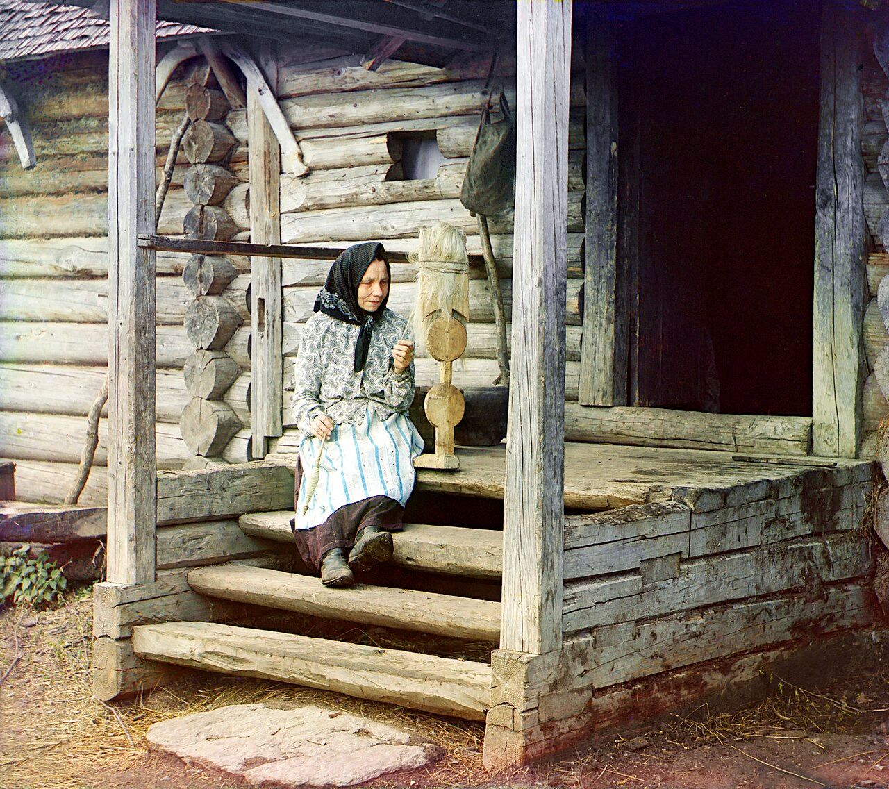 A woman in a dark headscarf, floral jacket and striped skirt sits on the wooden steps of a log house, a spindle in her hands and a tall carved distaff beside her.