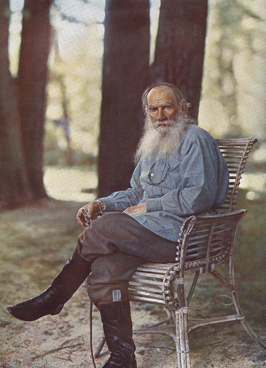 An elderly man with a long white beard, in a gray-blue shirt and tall black boots, sits in a wooden chair with his legs crossed among tall tree trunks.