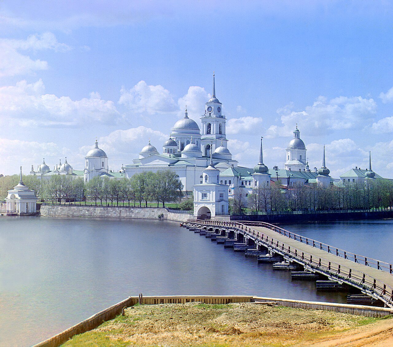 A white monastery with domes and a tall bell tower stands on an island across a calm lake, joined to the shore by a long wooden bridge, under a blue sky with white clouds.