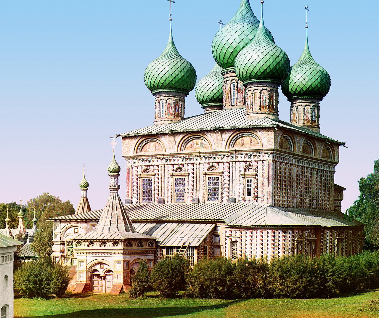 A church with patterned walls, a tiered roof and clustered green onion domes stands behind a hedge on a green lawn under a clear blue sky, a small bell tower at its left.