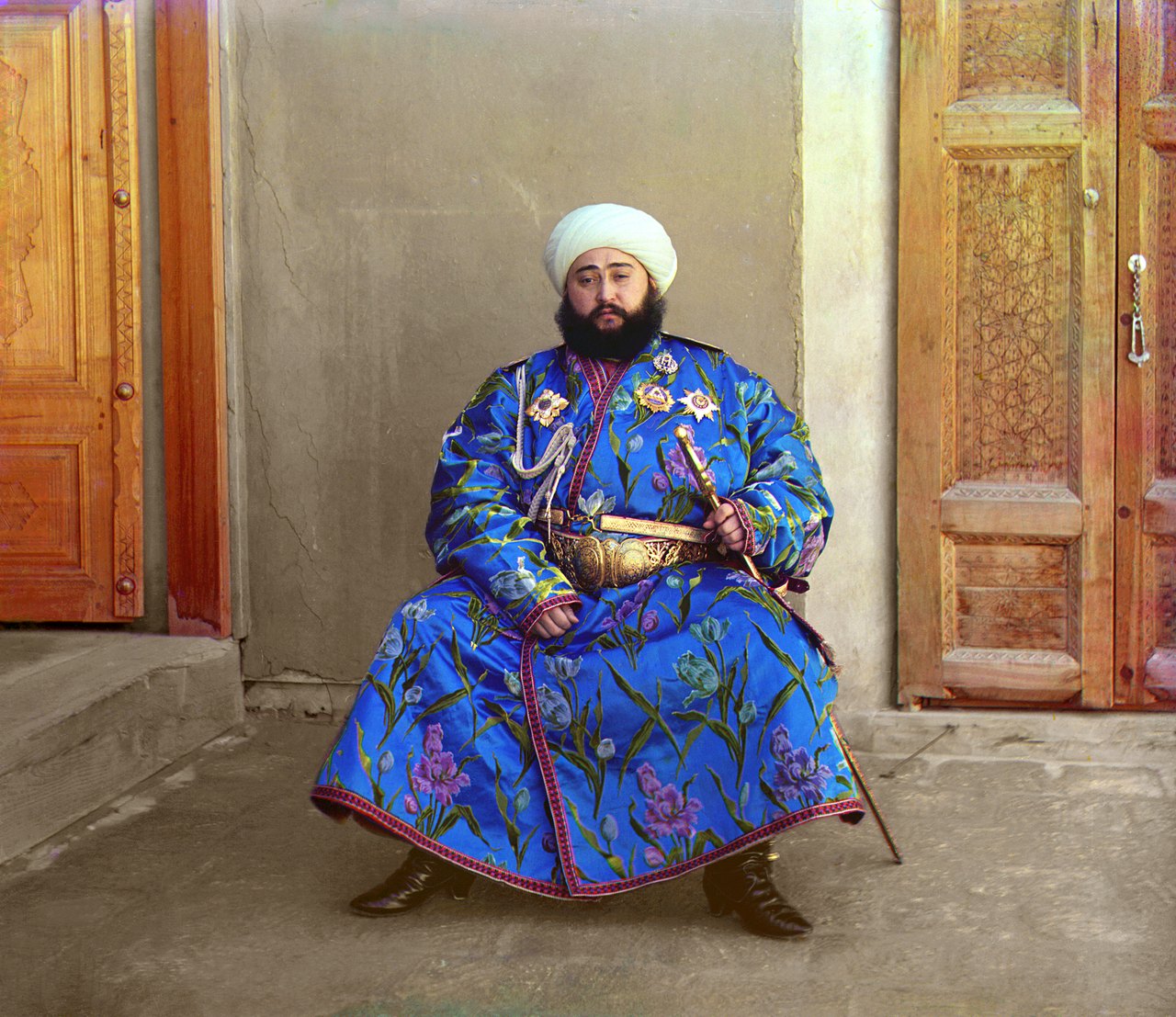 A bearded man in a white turban sits on a low seat in a blue robe patterned with flowers and pinned with medals, one hand on the hilt of a sword, between two carved wooden doors.