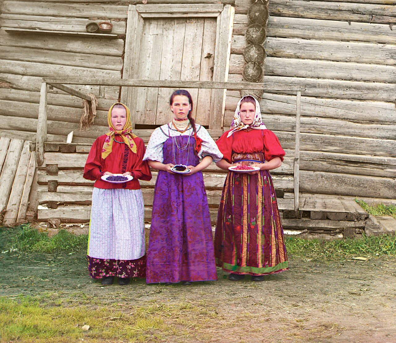 Three girls in bright red, purple and striped dresses stand in a row in front of a weathered log house, each holding a small plate of berries at waist height.