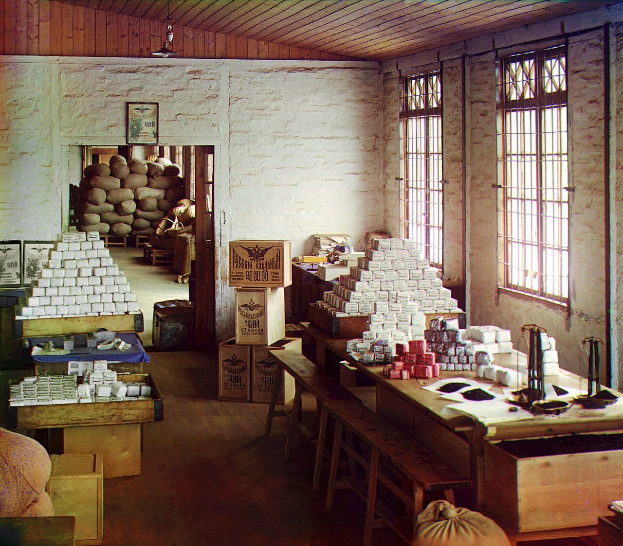 A bright, whitewashed room with tall windows holds long wooden tables piled with pyramids of white tea packages, a pair of scales and stamped crates, with stacked sacks in the room beyond.