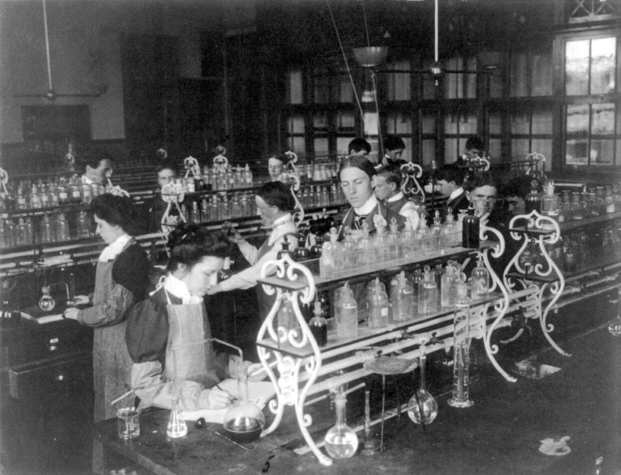 Students in aprons work at long laboratory benches crowded with glass bottles and flasks, with tall windows behind them.