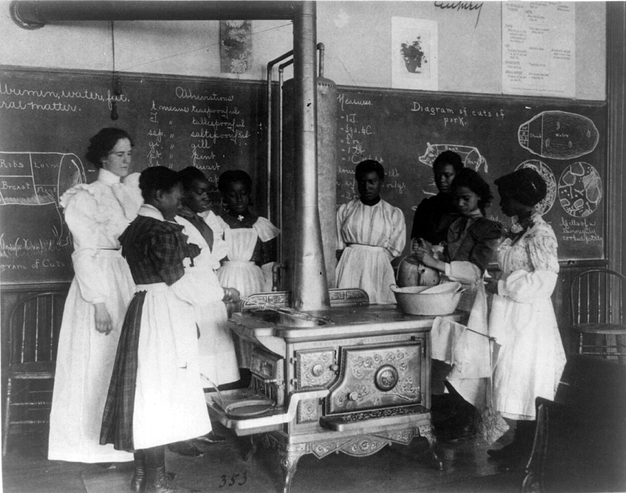 A teacher and a group of schoolgirls in white aprons stand around a wood stove in a classroom, with a blackboard of notes behind them.