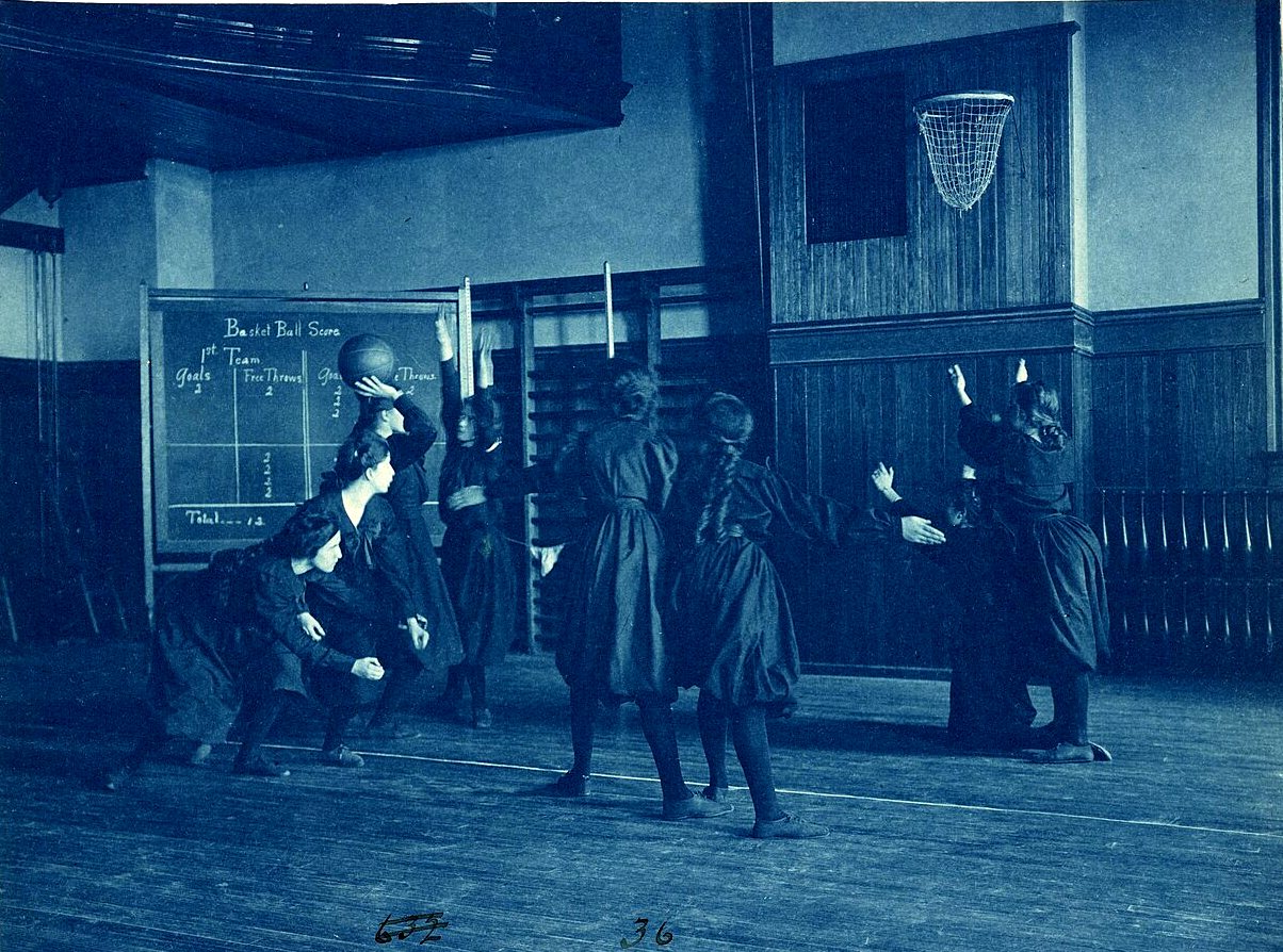 Girls in dark uniforms jump for a basketball under a hoop in a gymnasium, with a chalkboard scoreboard on the wall.