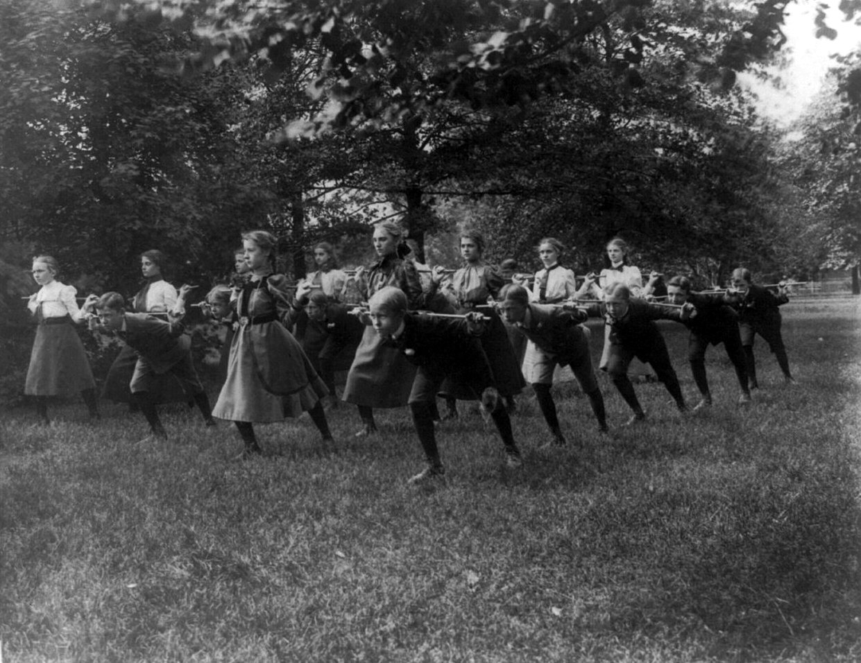 Children in a long line lean forward on a lawn under tall trees, each holding a rod out in front of them.