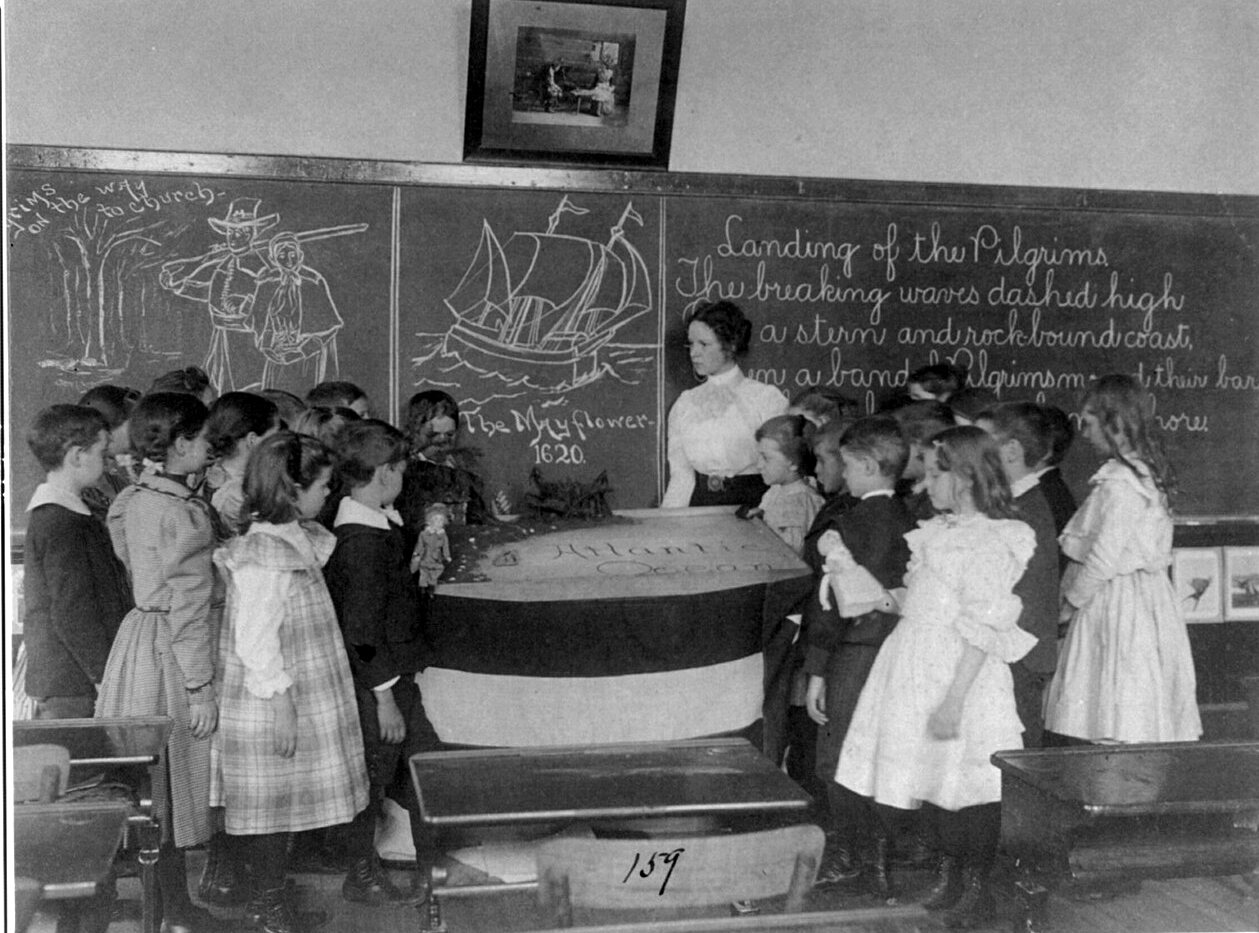Young children gather around a teacher at a blackboard covered with a chalk drawing of a sailing ship and a poem about the Pilgrims.