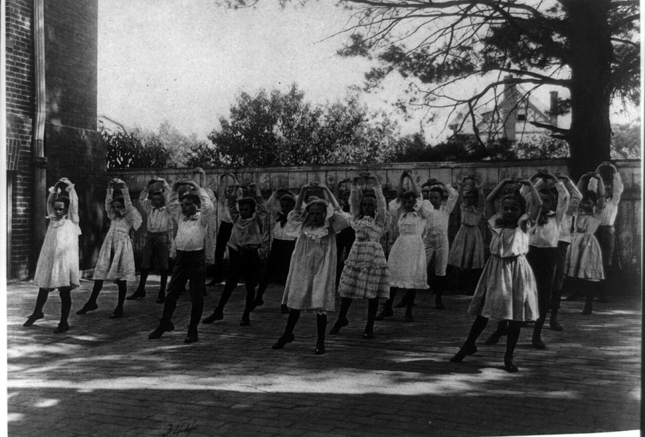 A line of children in light dresses and dark stockings dances with their hands behind their heads in a paved school yard.