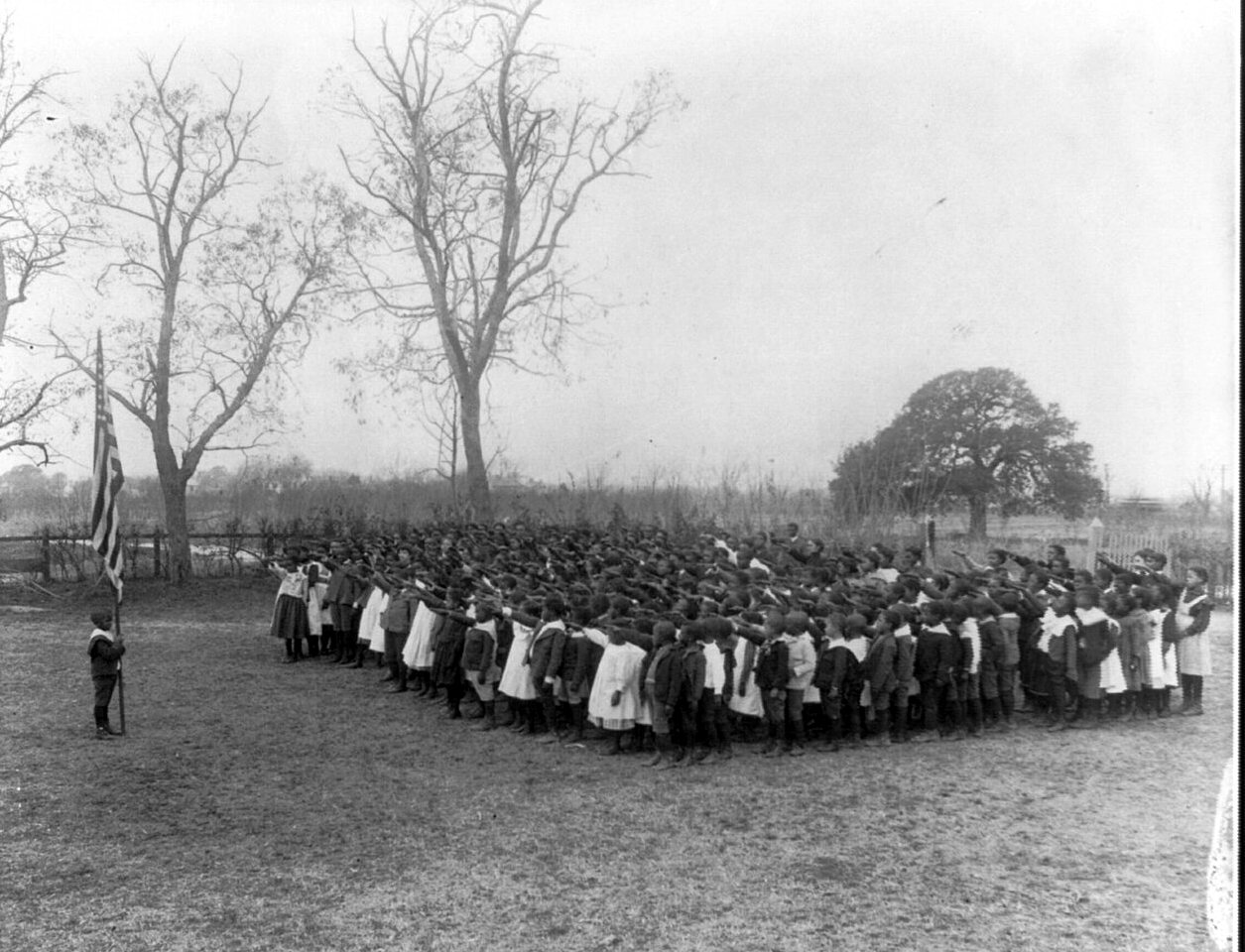 A large ring of schoolchildren stands on a bare field around a flag, with leafless trees behind.