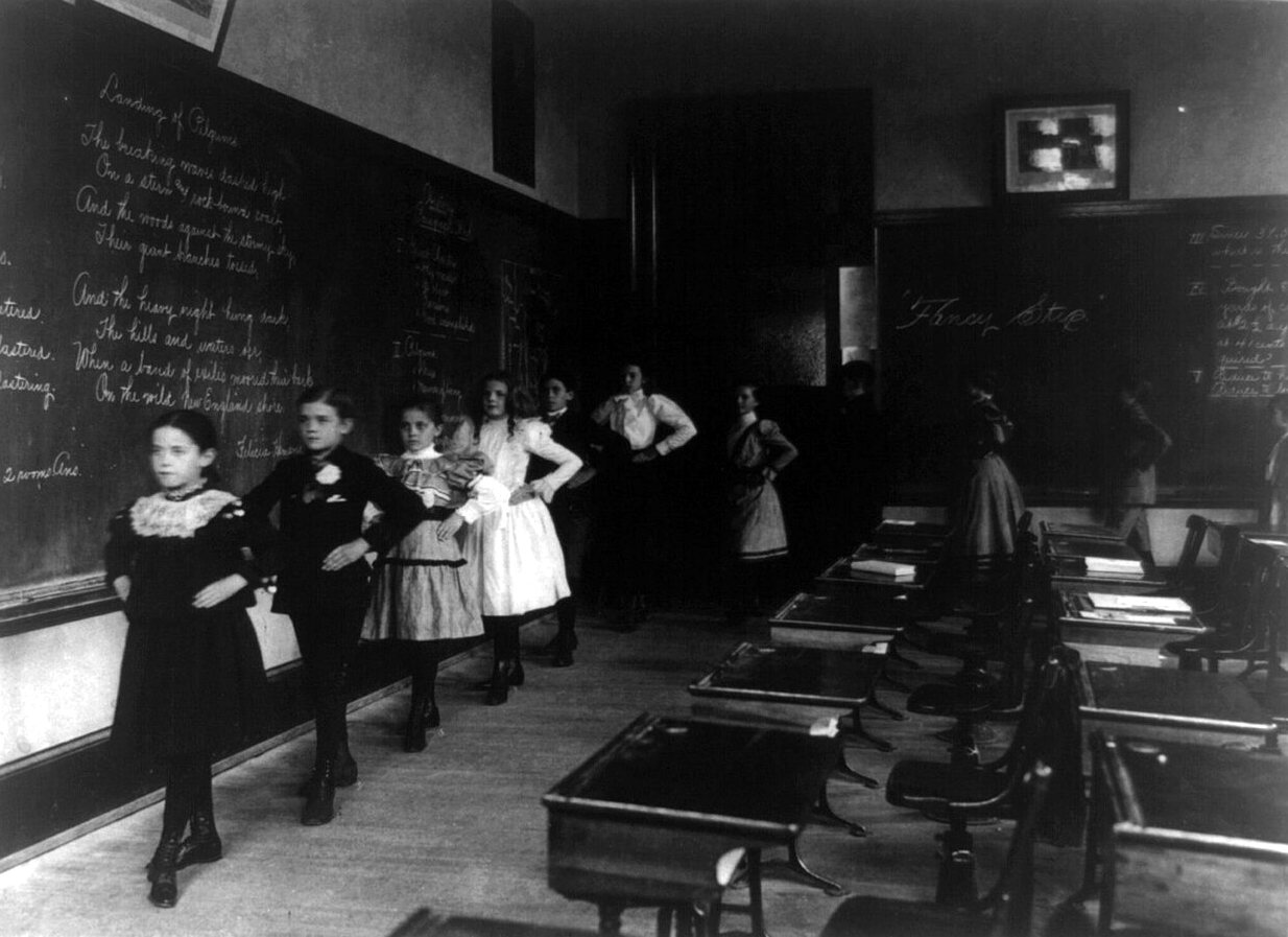 Girls in light and dark dresses walk in a line along the blackboards of a classroom, hands on their hips, beside rows of desks.