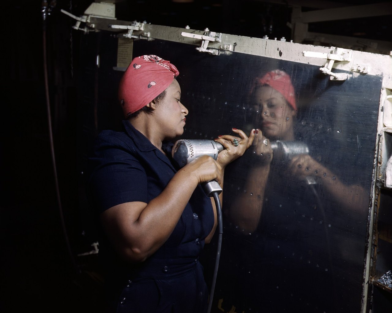 A woman in a red headscarf and a navy work shirt holds a hand drill against a dark, glossy panel, her face and the drill reflected in the surface at right.