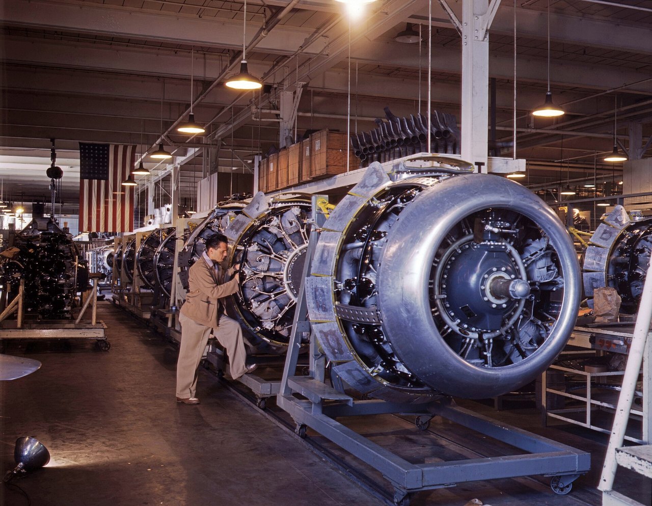 A line of silver radial engines on blue wheeled stands runs down a factory floor, with a man in tan coveralls working beside the second engine and an American flag hanging from the ceiling beyond.
