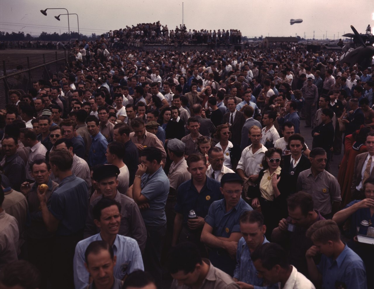 A dense crowd of workers, mostly men in work shirts and caps, fills the frame outdoors, with a smiling man in a light-blue shirt at the front, floodlight poles at upper left, a small balloon in the sky and an aircraft propeller at the far right edge.