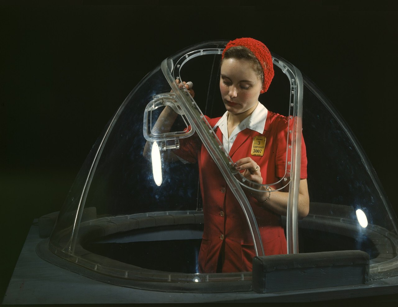 A woman in a red jacket with a white collar and a red knit cap stands inside a clear domed nose section, holding a curved strip of its transparent frame, a yellow badge pinned to her chest.