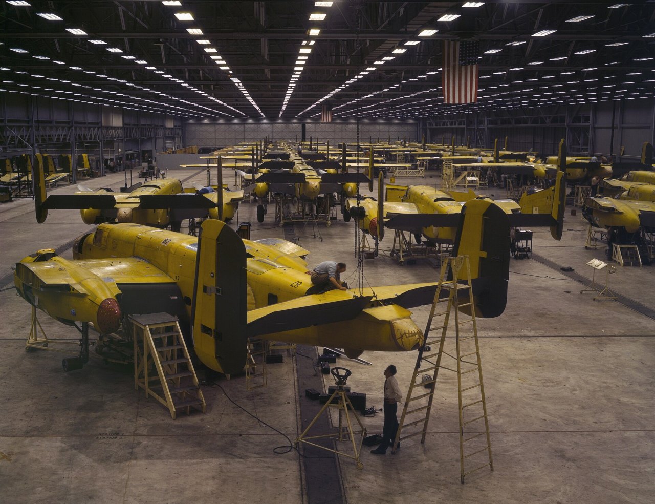 Rows of bright yellow bomber airframes stand in a huge factory hall under long lines of ceiling lights, with ladders and workers beside the nearest planes and an American flag hanging at the far end.