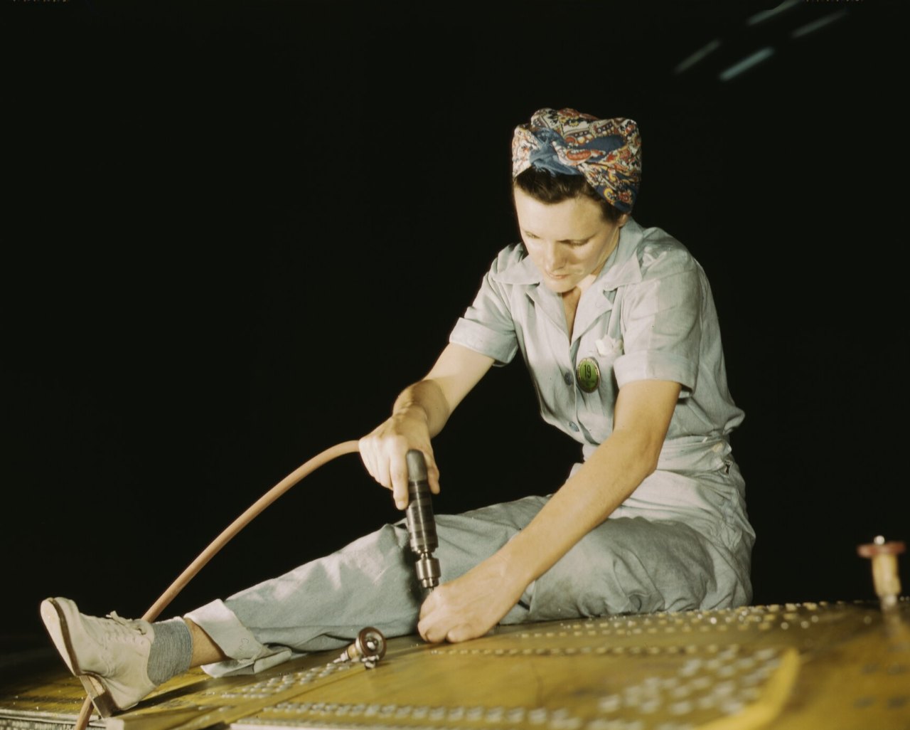 A woman in a patterned headscarf and pale gray-green coveralls sits on a metal surface dotted with rivet holes, drilling with a pneumatic drill, its hose looping around her boot, against a dark background.