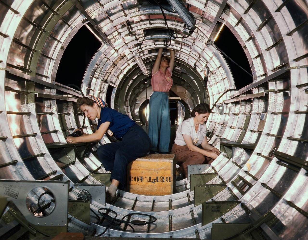 Three women work inside the ribbed, tunnel-like tail section of a bomber: one in blue overalls reaches left, one in a pink top and blue-green slacks stands on a wooden crate with her arms raised, and one in a white blouse works at right.