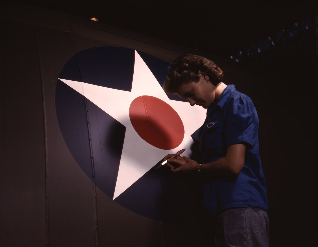 A woman in a blue work shirt leans toward the white star on a blue circle painted on a fuselage, a red dot at its center, touching it up with a small brush.