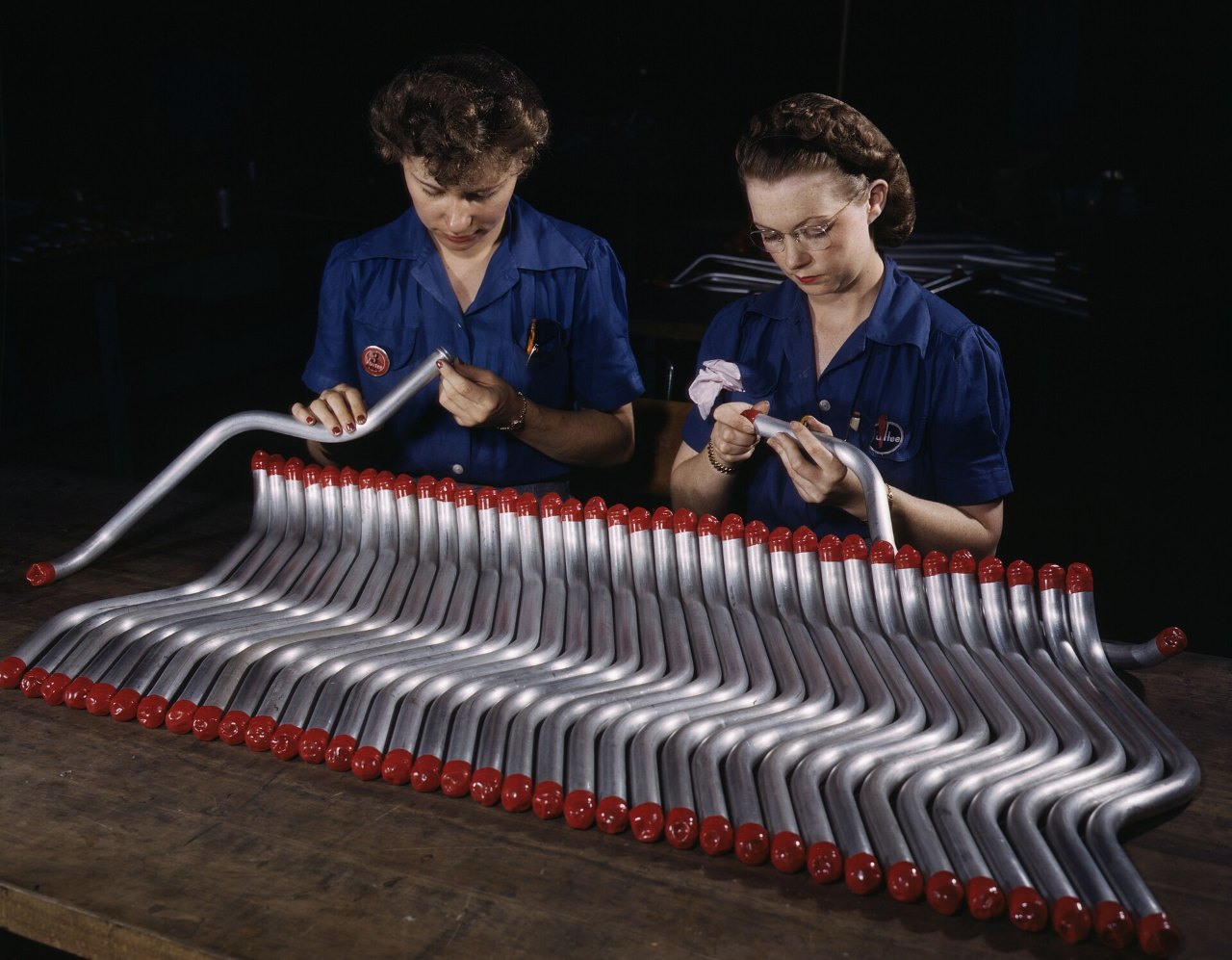 Two women in blue work shirts bend over a fan of bent silver tubes with red caps on their ends laid out on a wooden bench, one holding up a long tube and the other, in glasses, handling a short one, against a dark background.