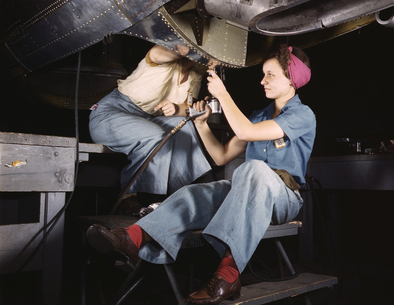 A woman in a pink headscarf and a light-blue shirt sits on a low platform and holds a pneumatic drill up against the underside of an aircraft, while a second woman in a cream blouse works behind her.